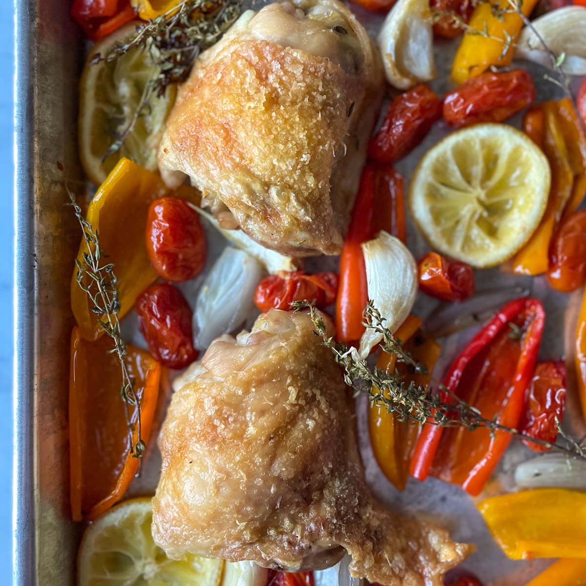 Two crispy skinned chicken thighs sit on a sheet tray surrounded by roasted baby sweet peppers, sliced lemon, quartered shallots, garlic, thyme, and cherry tomatoes.