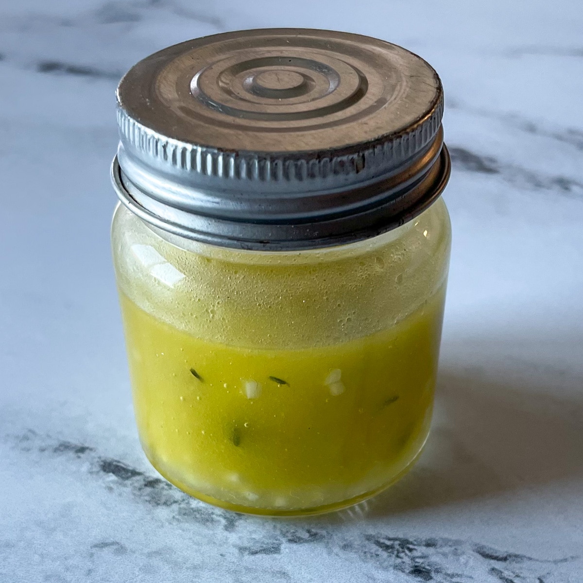 A small clear glass jar with a silver lid containing lemon, garlic, thyme vinaigrette sits on a marble counter top.