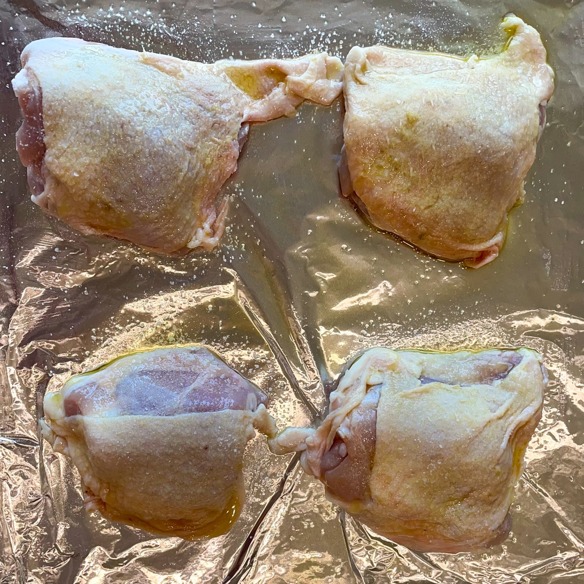 Salted bone-in, skin-on chicken thighs rubbed with olive oil sit on a sheet tray coated with a bit of olive oil.