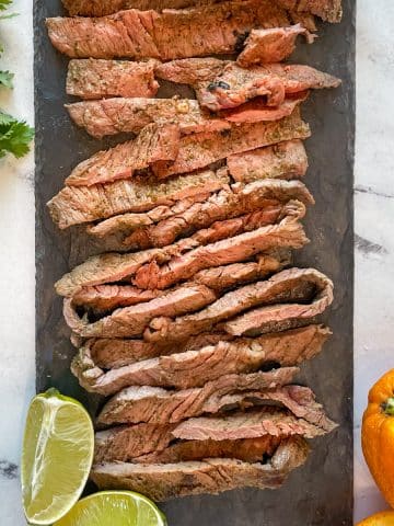 Grilled, sliced ranchera meat is shown on a black stone plate surrounded by lime and orange slices and cilantro.