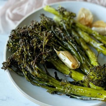 Roasted tenderstem broccoli and garlic are shown on a white plate with a light pink linen in the background and a silver fork in the foreground.