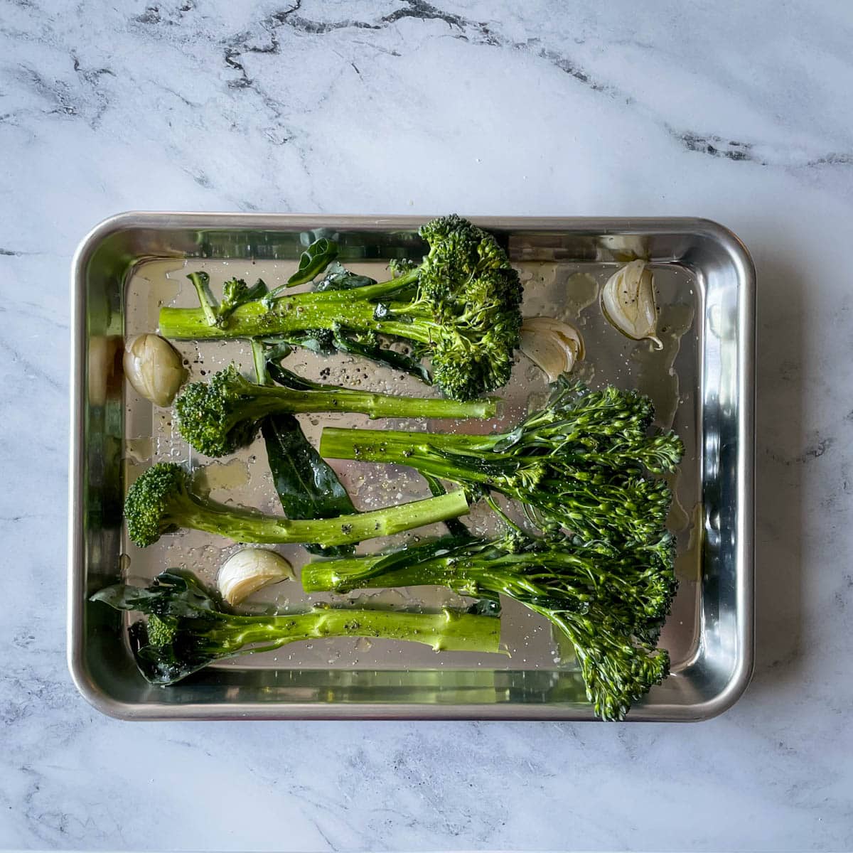 Tenderstem broccoli and garlic cloves in their skin sit on a quarter sheet pan drizzled with olive oil, salt, and pepper.