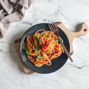 Twirled spaghetti arrabbiata on a black plate with a copper fork topped with parsley on a wooden cutting board over a white marble counter top with a gray linen in the background.