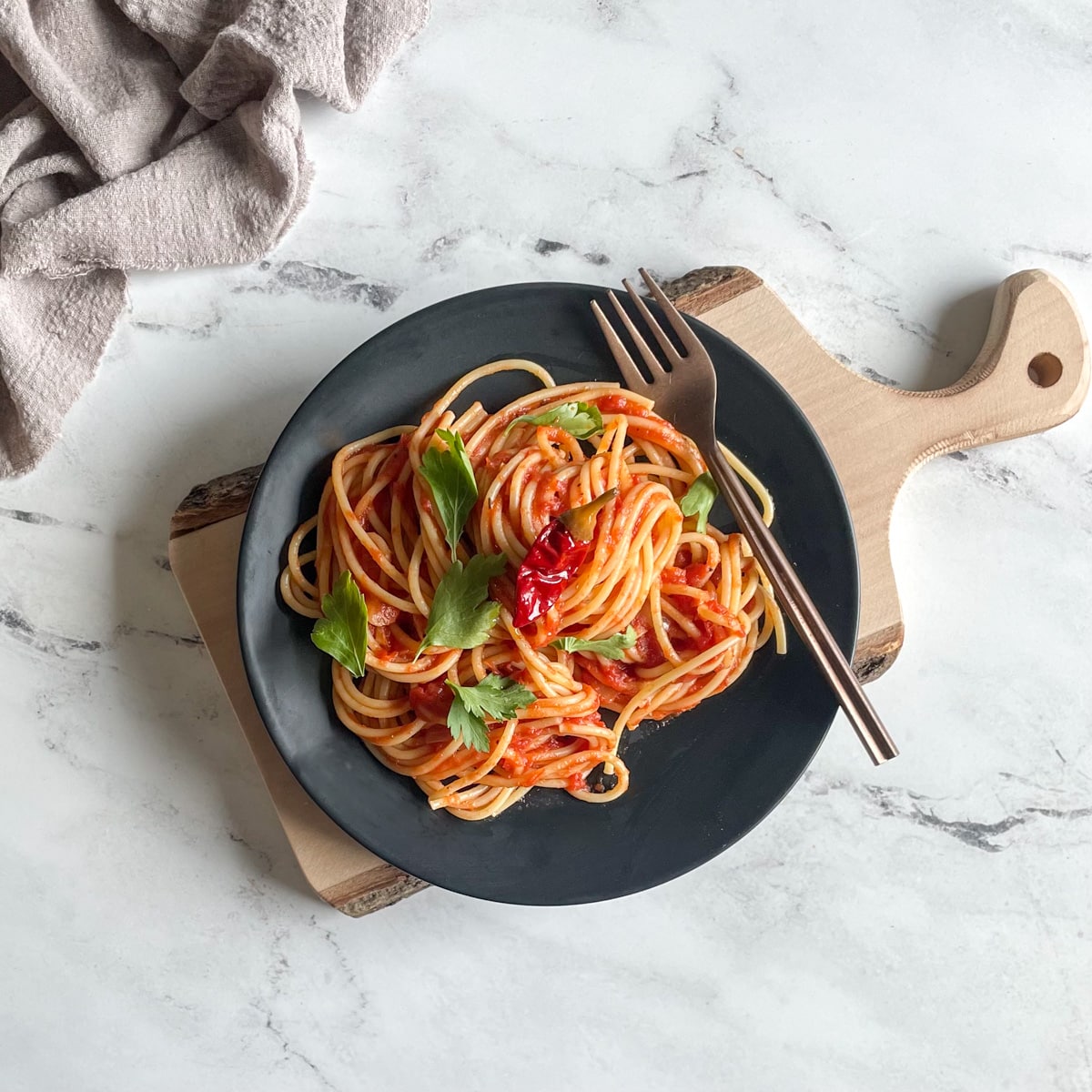 Twirled spaghetti arrabbiata on a black plate with a copper fork topped with parsley on a wooden cutting board over a white marble counter top with a gray linen in the background.