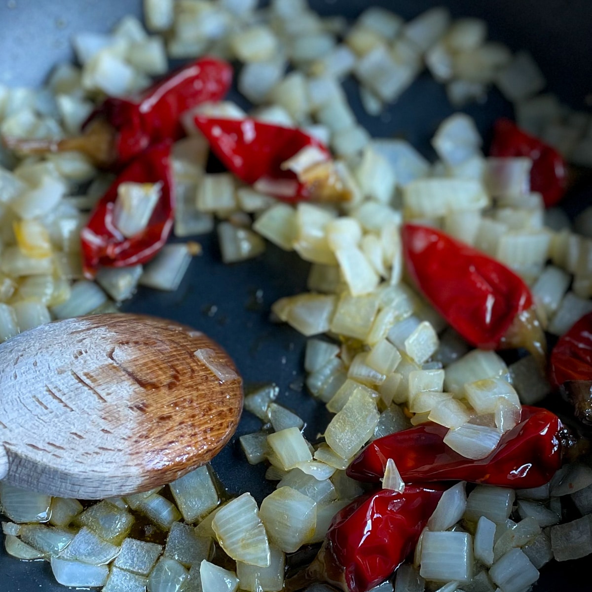 Sweating onions and Calabrian chilis are mixed with a wooden spoon in a black pan.