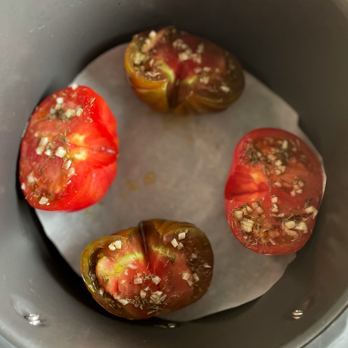 One halved heirloom red tomato and one halved purple and red heirloom tomato coated in oil, spices, and minced garlic sit on a piece of parchment paper in an air fryer basket.