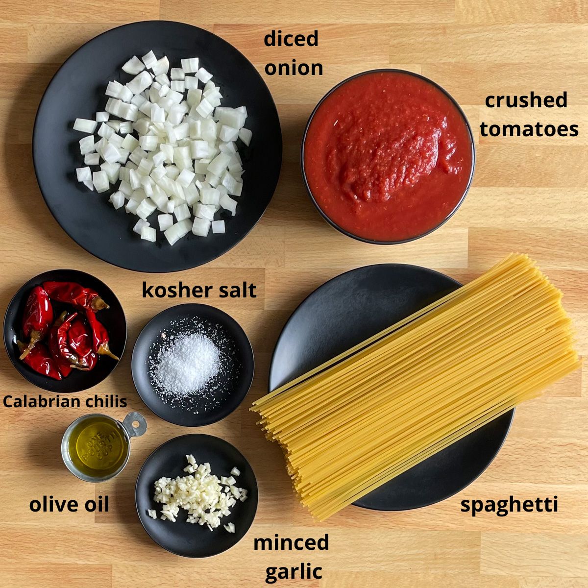 Diced onion, crushed tomatoes, Calabrian chilis, kosher salt, olive oil, minced garlic, and spaghetti sit on black plates on a butcher block cutting board. Each item is labeled in black print.