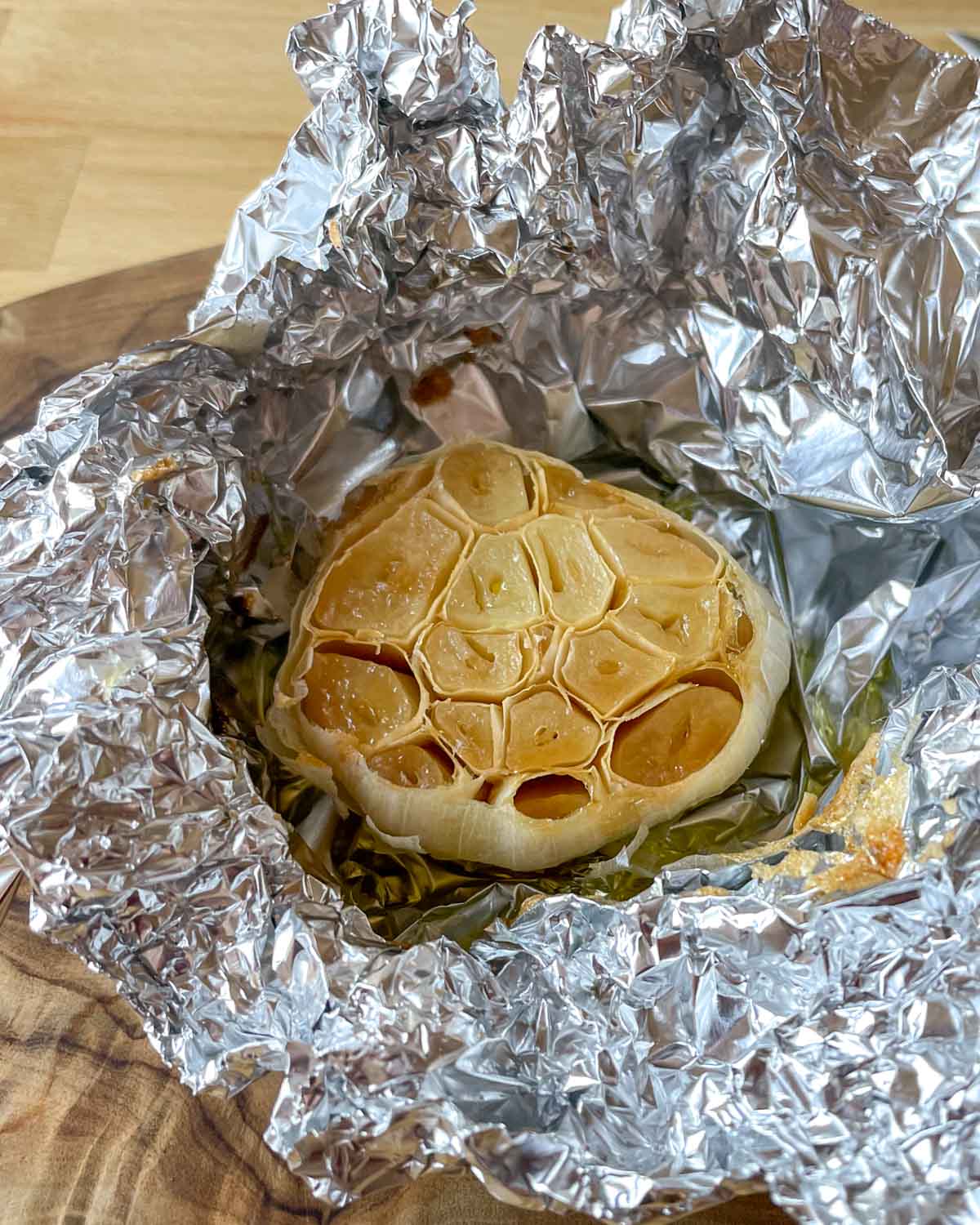 A head of roasted garlic sits in tin foil on a wooden cutting board.