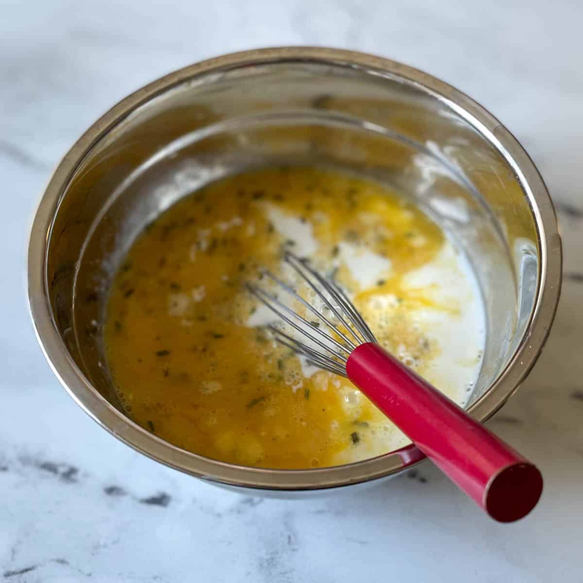 Beaten egg, milk, and chopped rosemary are shown in a metal bowl with a red whisk.