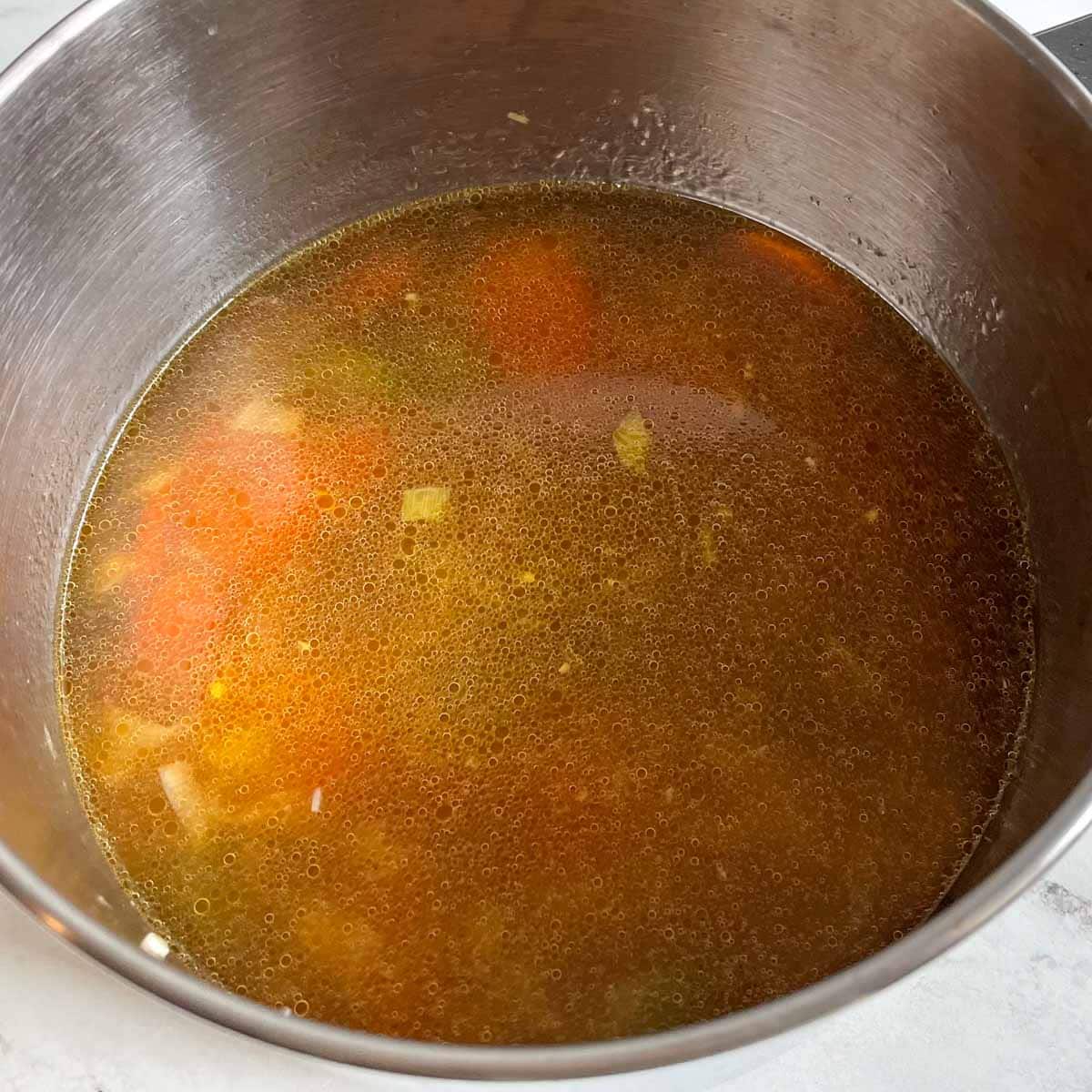 Vegetable broth with sliced carrots, celery, and leek are shown in a stainless steel pot.
