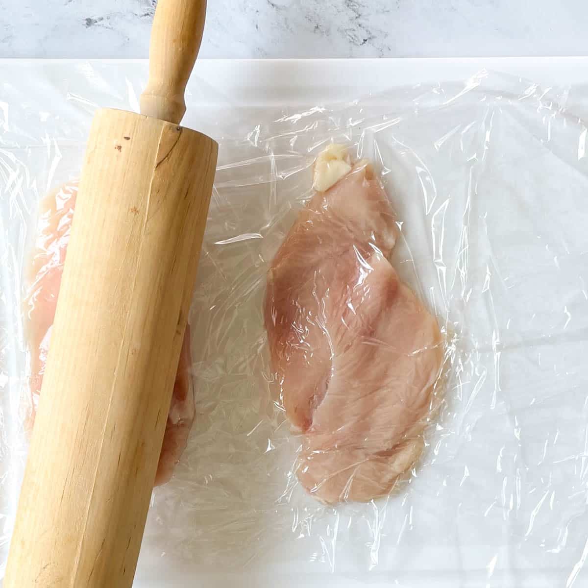 Two cutlets are showing on a white cutting board covered with plastic wrap with a rolling pin flattening the cutlets.