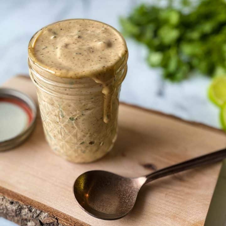 Chipotle Sauce is shown in a glass jar on a wooden cutting board with a copper spoon and parsley and lime wedges.