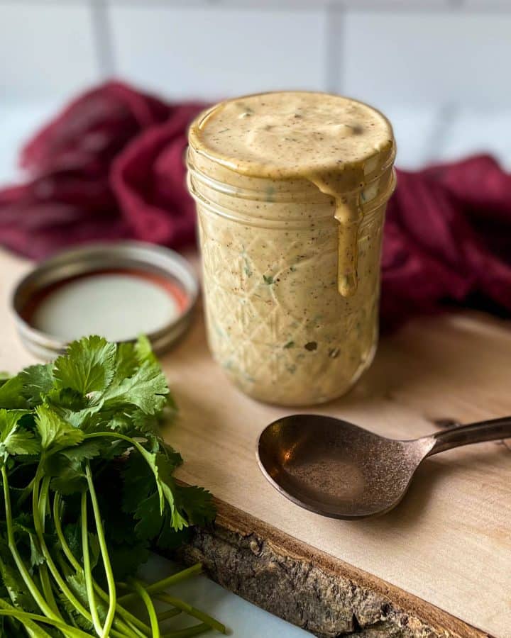 Chipotle sauce in a glass jar sits on a wooden cutting board with a spoon, parsley and a red linen.