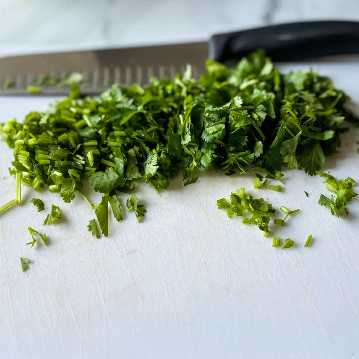 Chopped fresh cilantro is shown on a white cutting board with a kitchen knife in the background.
