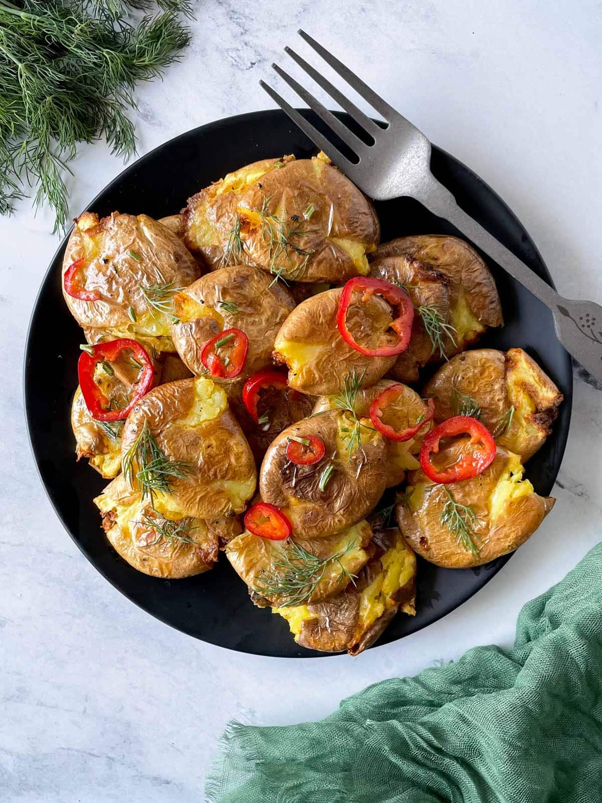 Smashed air fryer potatoes are shown dressed in lemon, rosemary, chili vinaigrette on a black plate with a green linen and a bunch of dill.