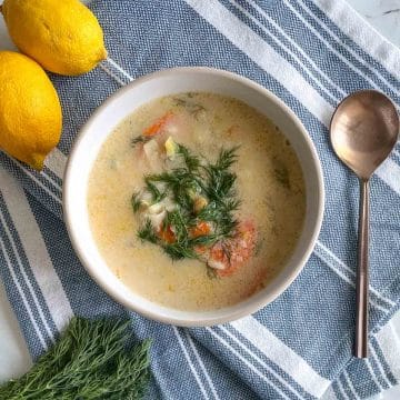 A bowl of Greek Chickpea Soup garnished with dill sits on a blue and white linen with a spoon, fresh dill, and lemons.