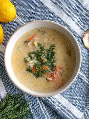 A bowl of Greek Chickpea Soup garnished with dill sits on a blue and white linen with a spoon, fresh dill, and lemons.
