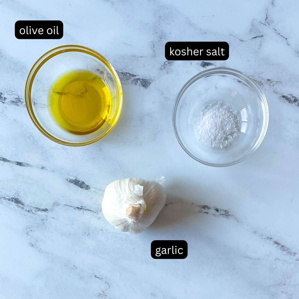 Olive oil, kosher salt, and a head of garlic are shown on a white marble counter.