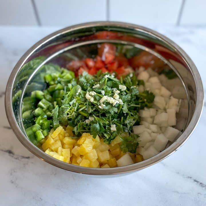 The chopped ingredients for pineapple pico de gallo sit in a stainless steel bowl topped with minced garlic.