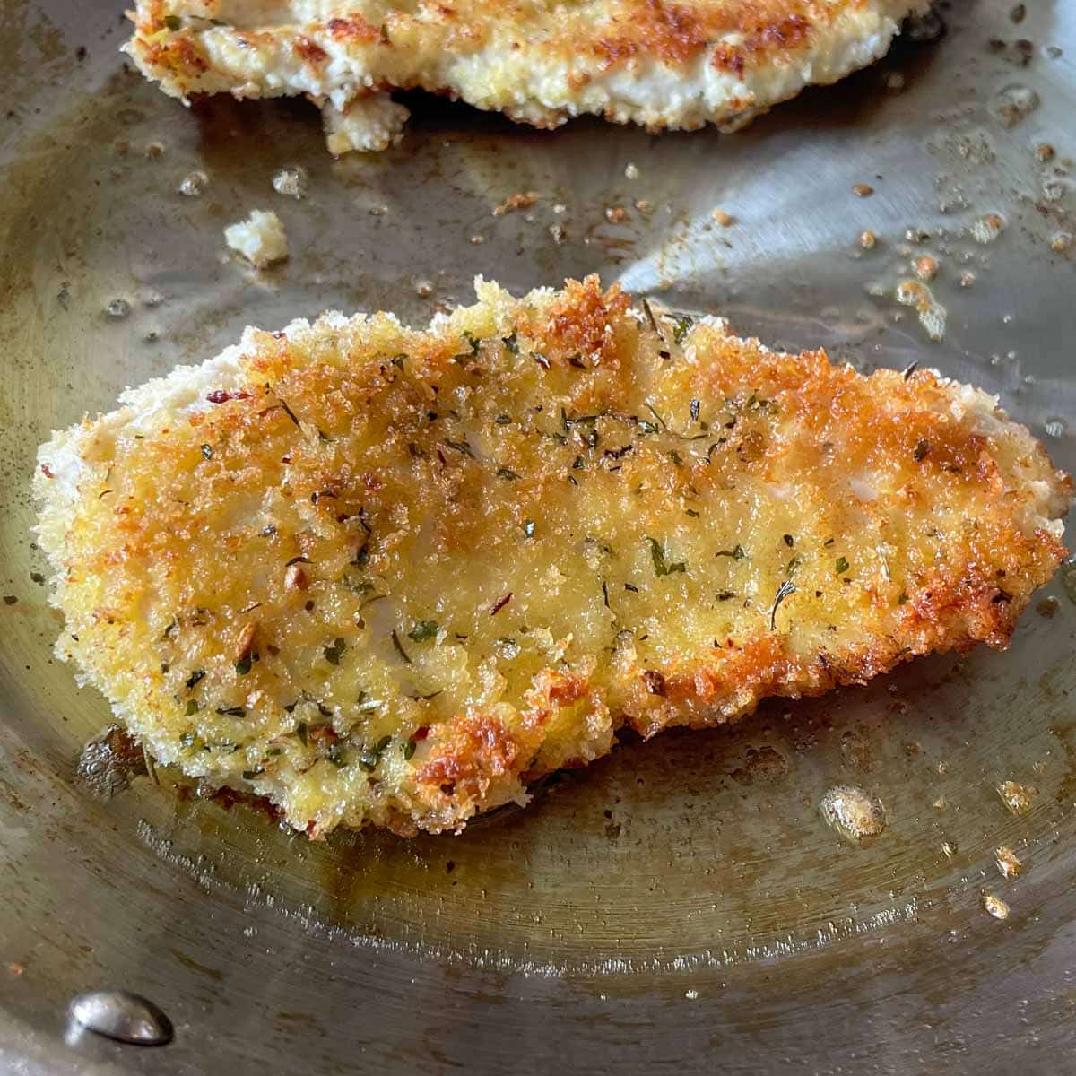 Fried, breaded chicken cutlet cooking in a stainless steel frying pan.