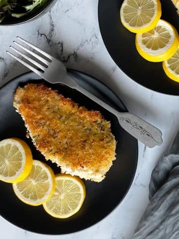 A fried, breaded chicken cutlet with three lemon slices on a black plate on a white marble counter.