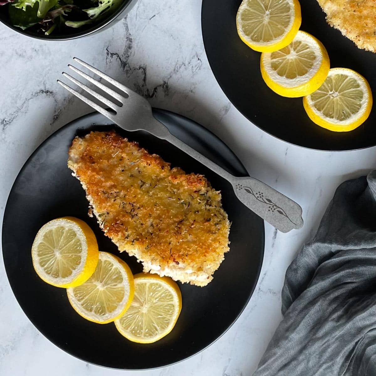 A fried, breaded chicken cutlet with three lemon slices on a black plate on a white marble counter.