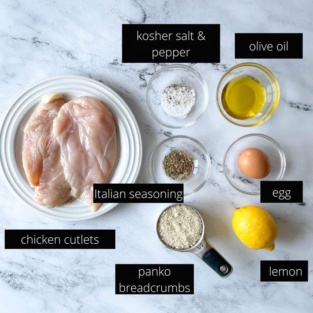 The labeled ingredients for Italian Chicken Cutlets sit on a white marble counter; they include: chicken cutlets, olive oil, kosher salt, pepper, Italian seasoning, an egg, a lemon, and panko breadcrumbs.