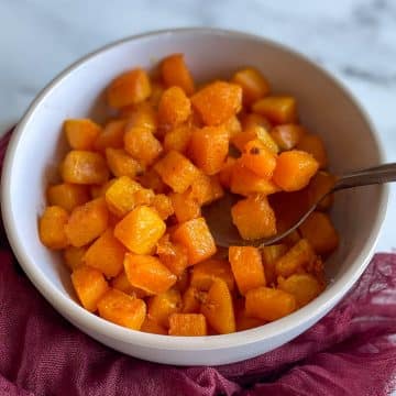 Sautéed butternut squash is shown in a white bowl with a red linen and a copper spoon on a white marble counter.