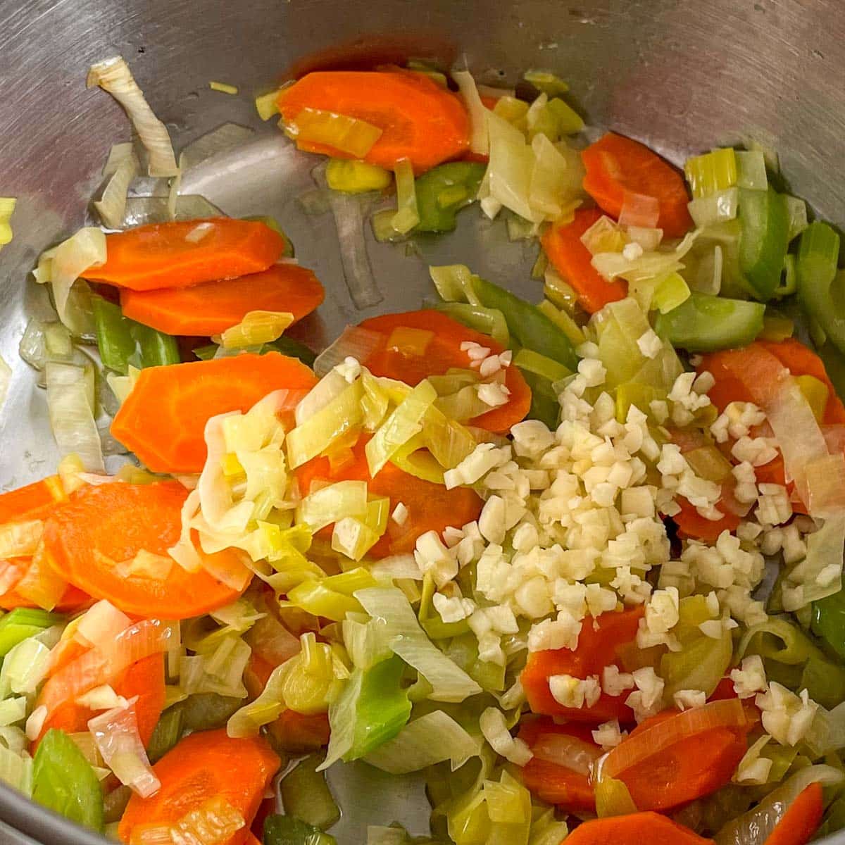 Minced garlic is added to sauteed carrot, celery, and leek in a stainless steel pot.