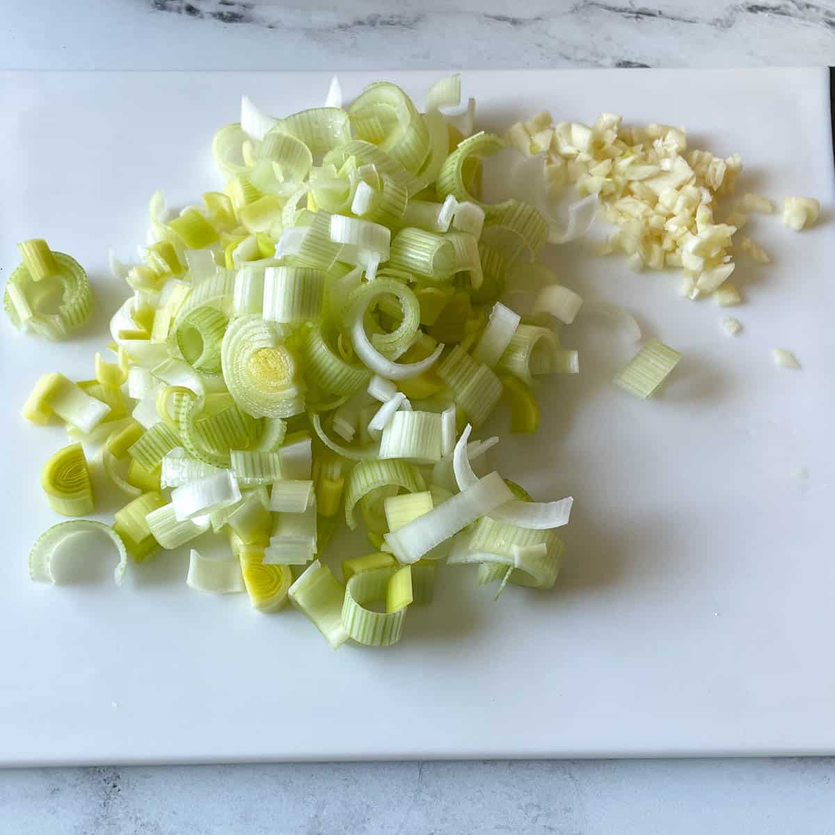 sliced leek and minced garlic sit on a white cutting board.