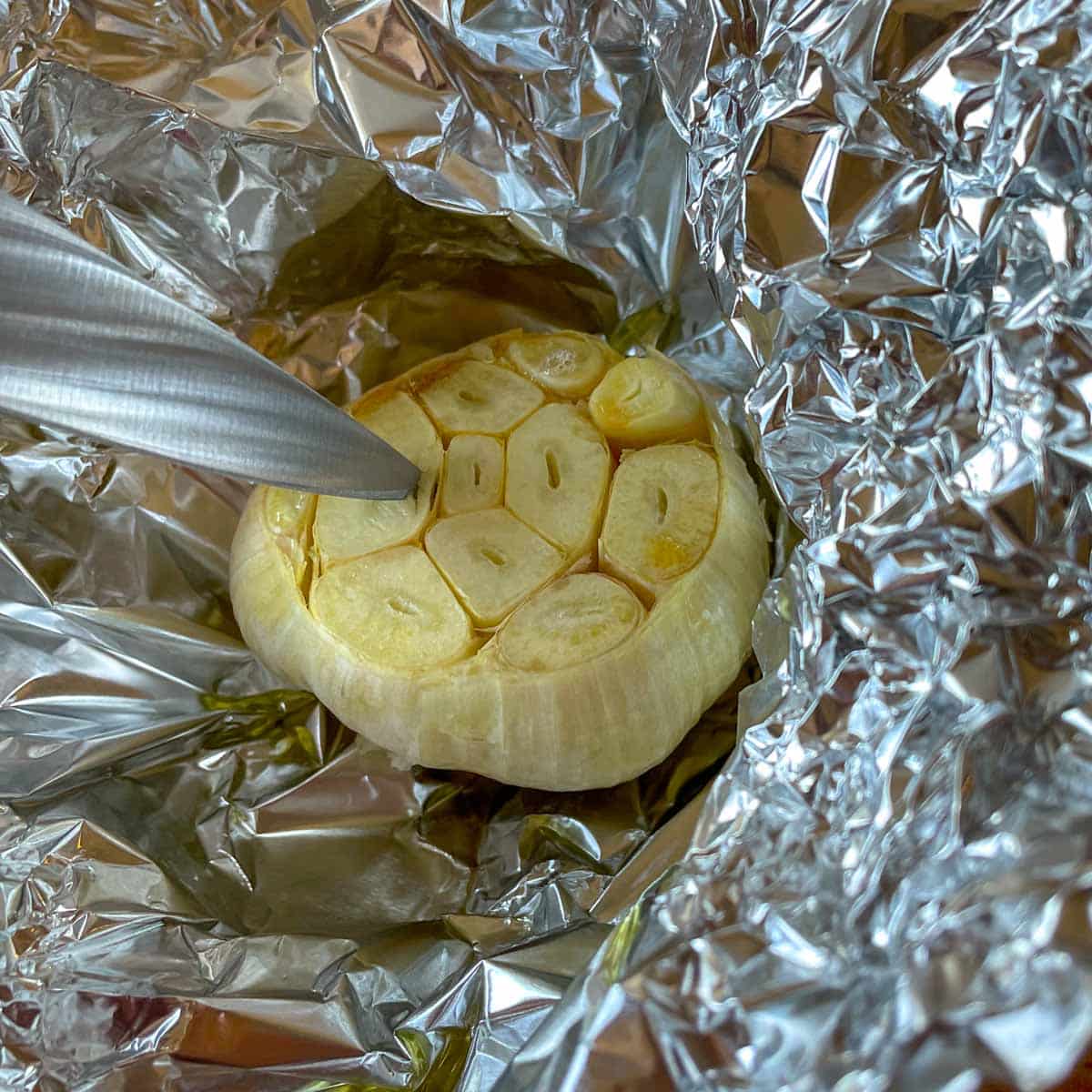 Partially roasted garlic is shown in tin foil; a paring knife is inserted into one of the cloves to test doneness.