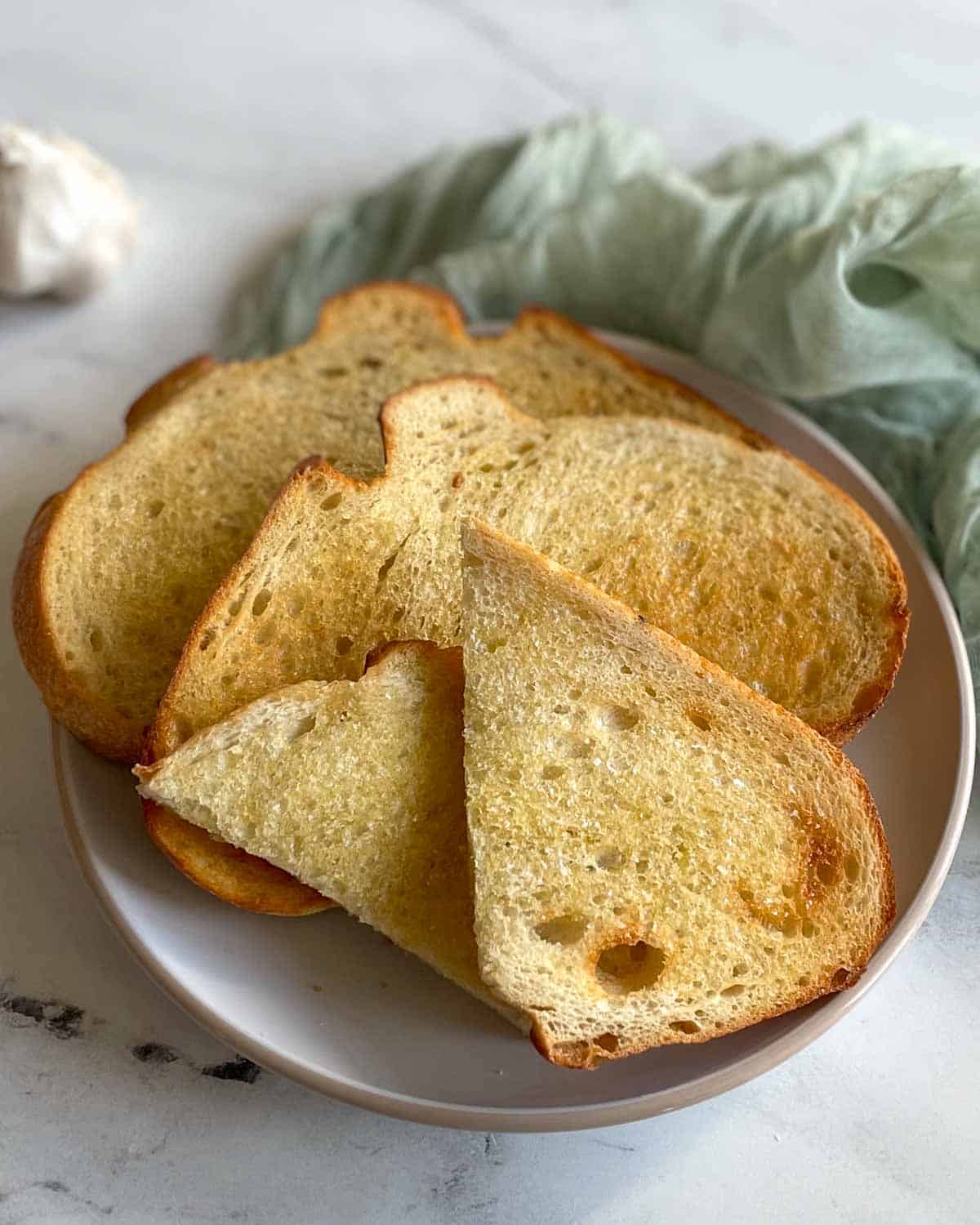 Three slices of toasted bread are shown on a white plate on a white marble counter with a light green linen.