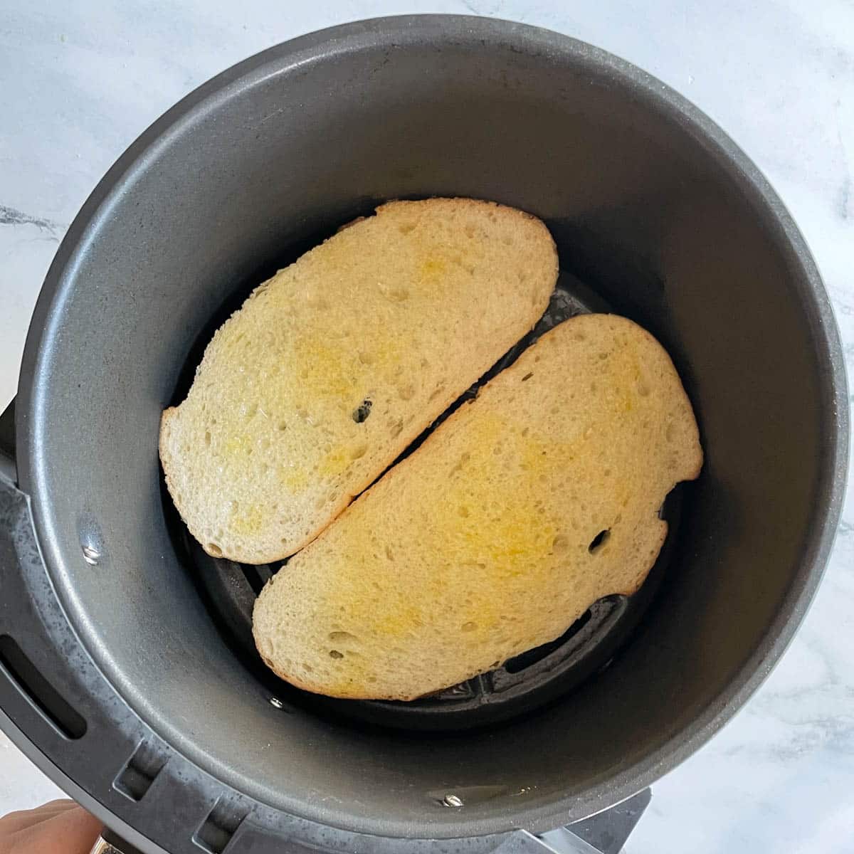 Two slices of bread brushed with olive oil are shown in the basket of an air fryer.