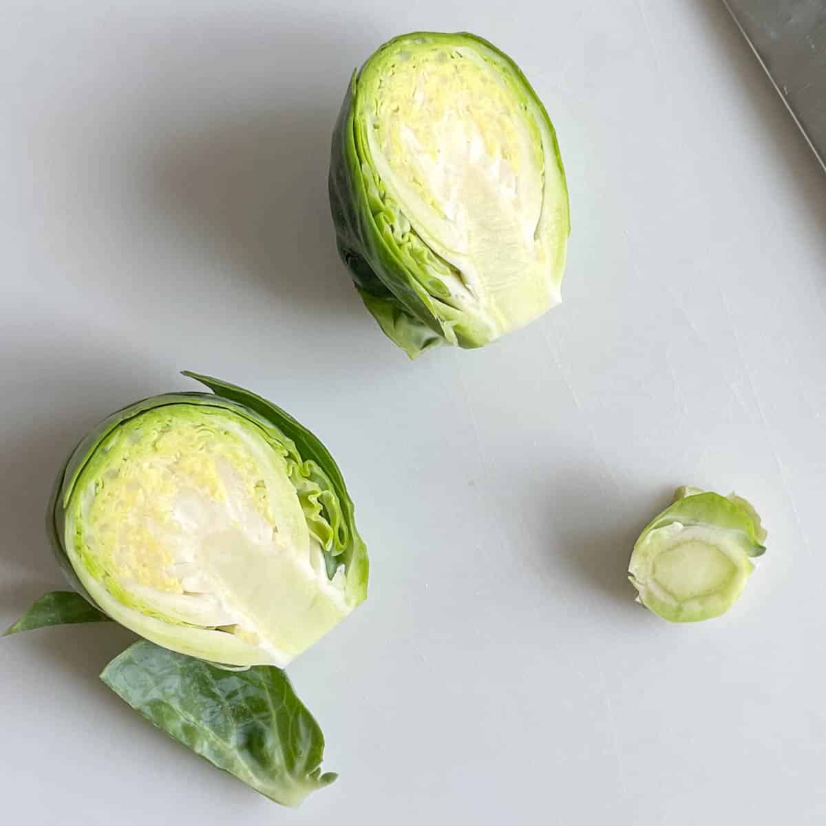 A halved brussels sprout with the end removed are shown on a white cutting board.