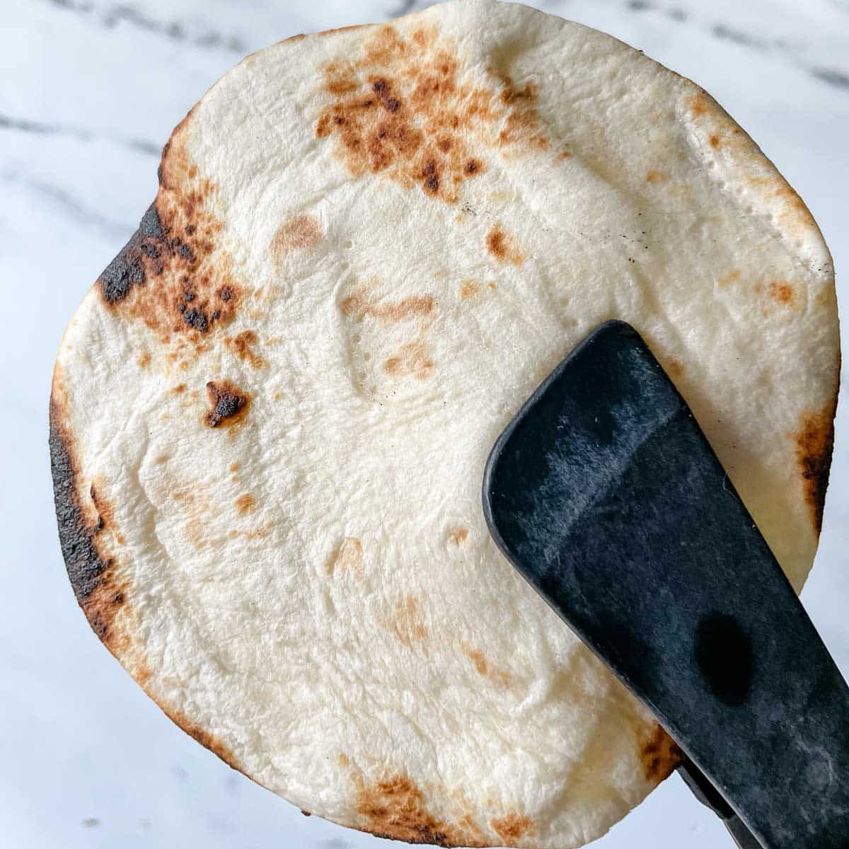 A pair of tongs holds up a lightly charred tortilla.