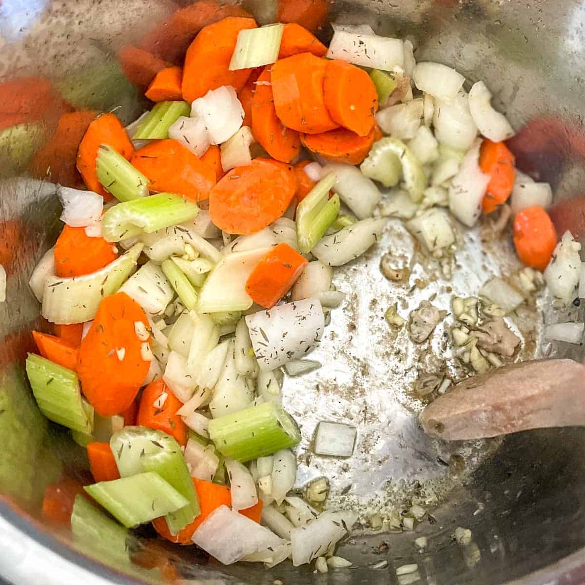 Sliced carrots, celery, chopped onions and garlic, olive oil, and thyme are shown in the instant pot, stirred with a wooden spoon.