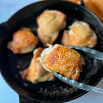 A golden brown chicken thigh is held with tongs in the foreground above a black cast iron pan of chicken thighs.