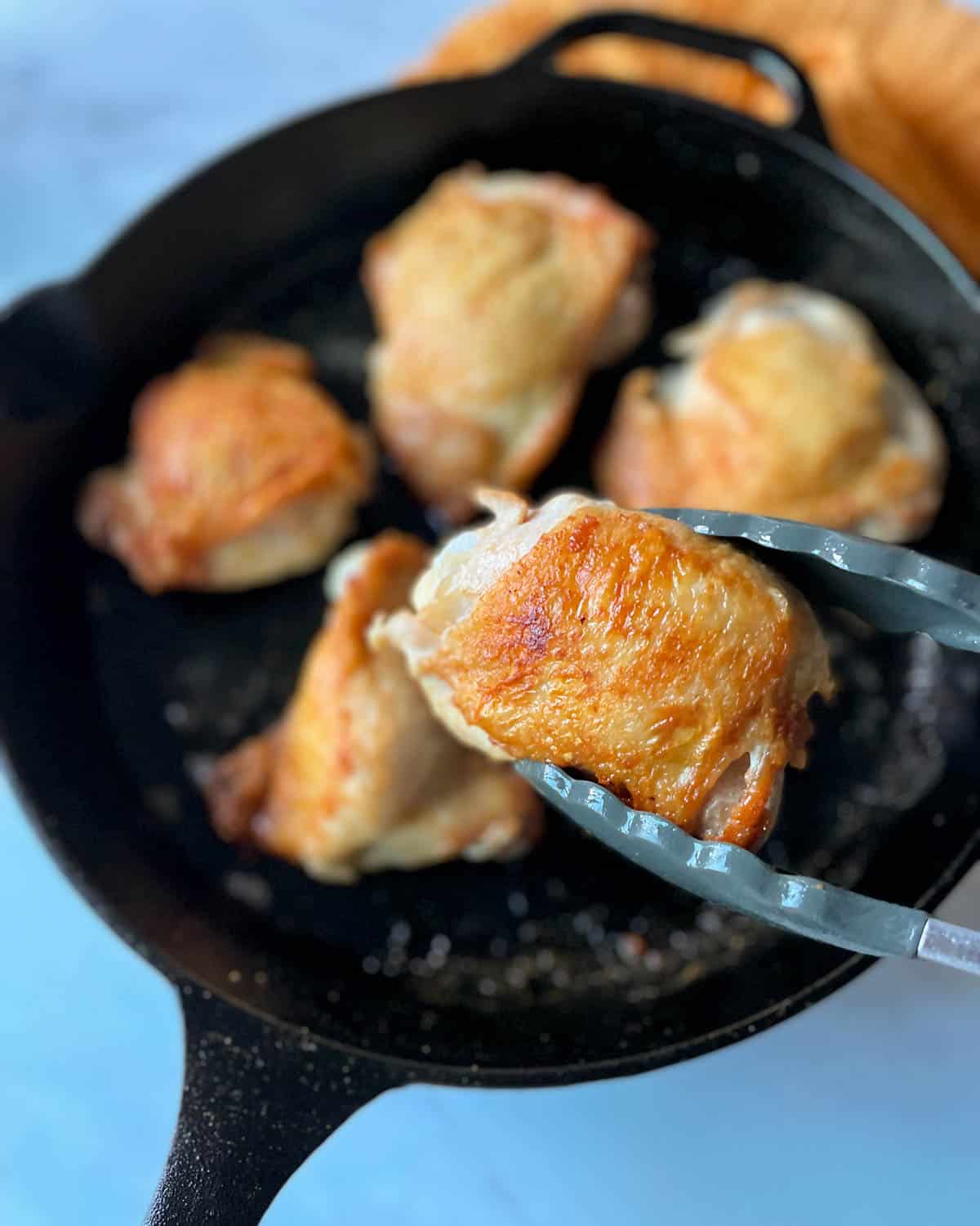 A golden brown chicken thigh is held with tongs in the foreground above a black cast iron pan of chicken thighs.