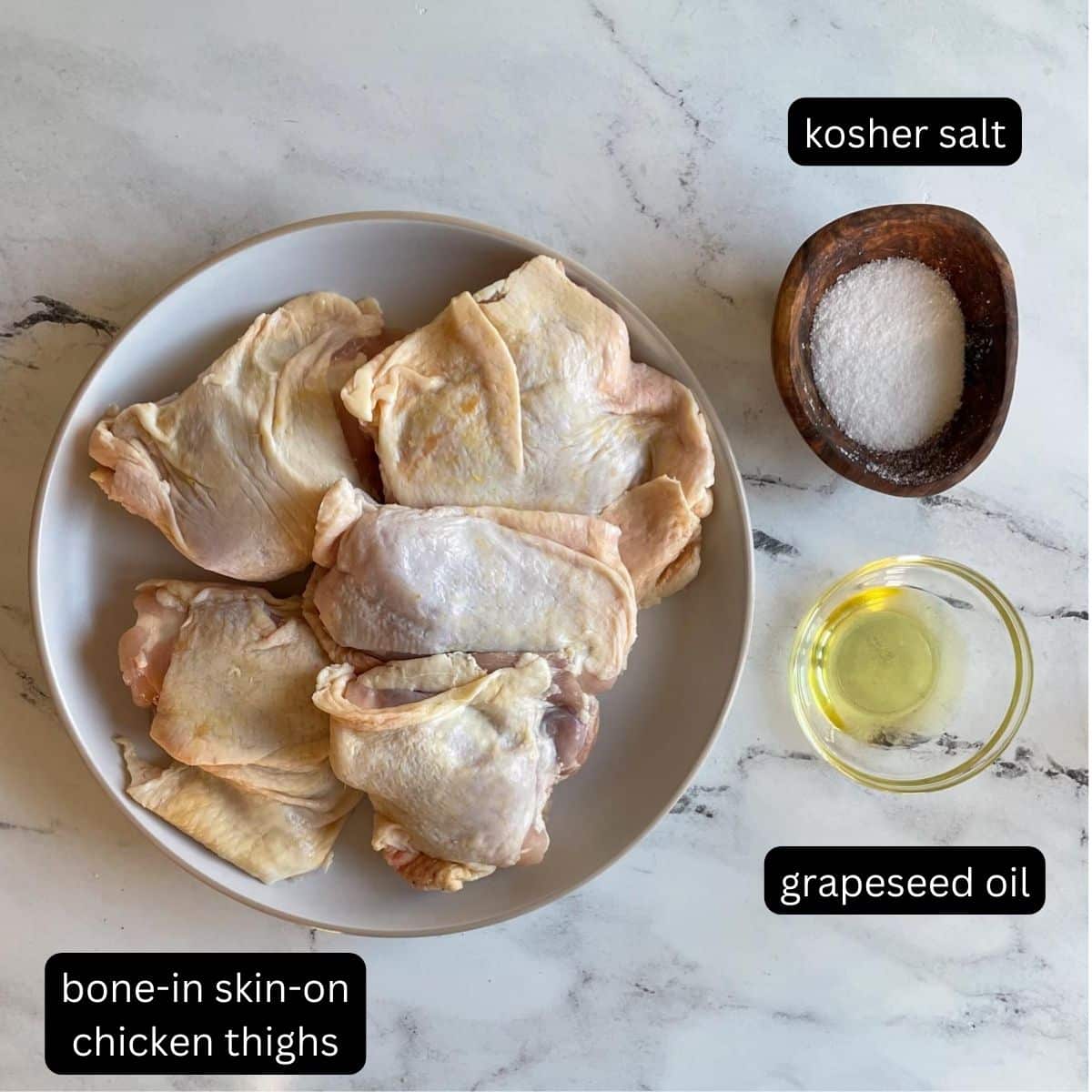 The labeled ingredients for Cast Iron Chicken sit on a white marble counter; they include: bone-in, skin-on chicken thighs, grapeseed oil, and kosher salt.