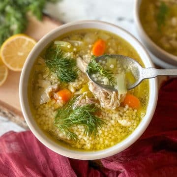 A bowl of chicken pastina soup sits on a rustic wooden cutting board with lemons and dill in the background.