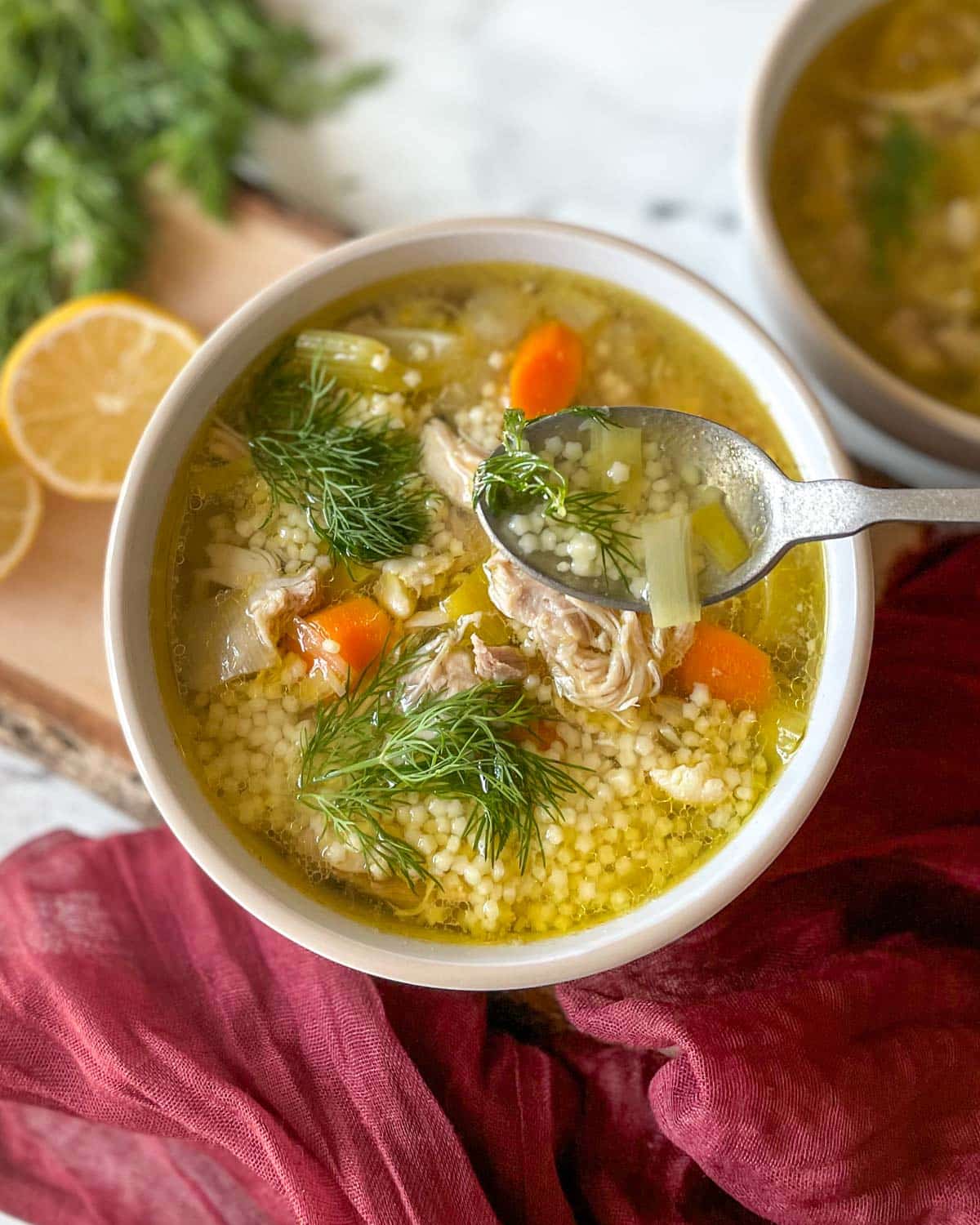 A bowl of chicken pastina soup sits on a rustic wooden cutting board with lemons and dill in the background.
