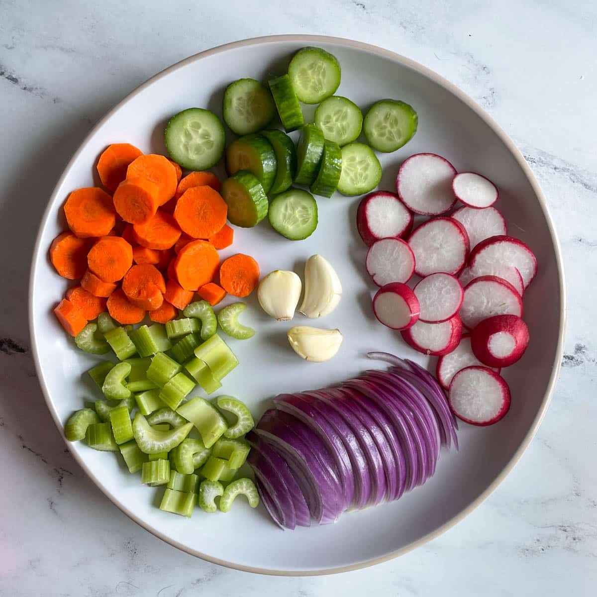 Sliced red onion, celery, carrot, cucmber, radish, and smashed garlic cloves sit on a white plate.