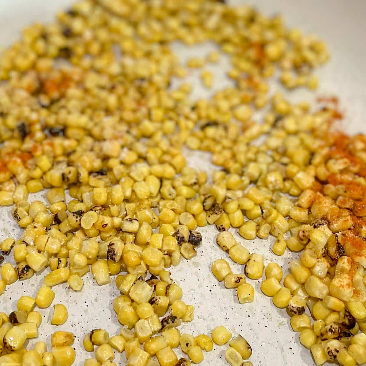 Roasted corn kernels dusted with garlic powder, cayenne, and salt are sautéed in a white frying pan.