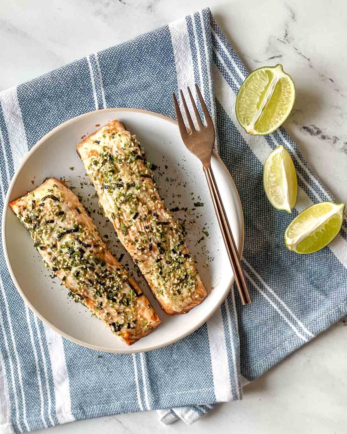 Two baked salmon fillets coated in mayonnaise and furikake sit on a white plate on a blue and white linen with lime slices.