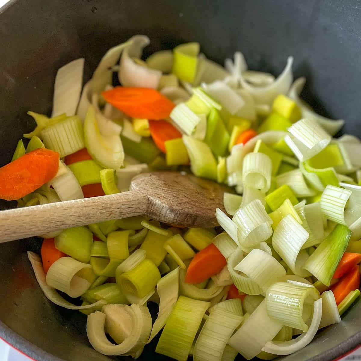 Leeks, celery, and carrots are stirred with wooden spoon in a black cast iron pot.