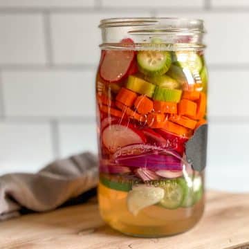 A glass jar of mixed pickles sits on a wooden cutting board with a gray linen.
