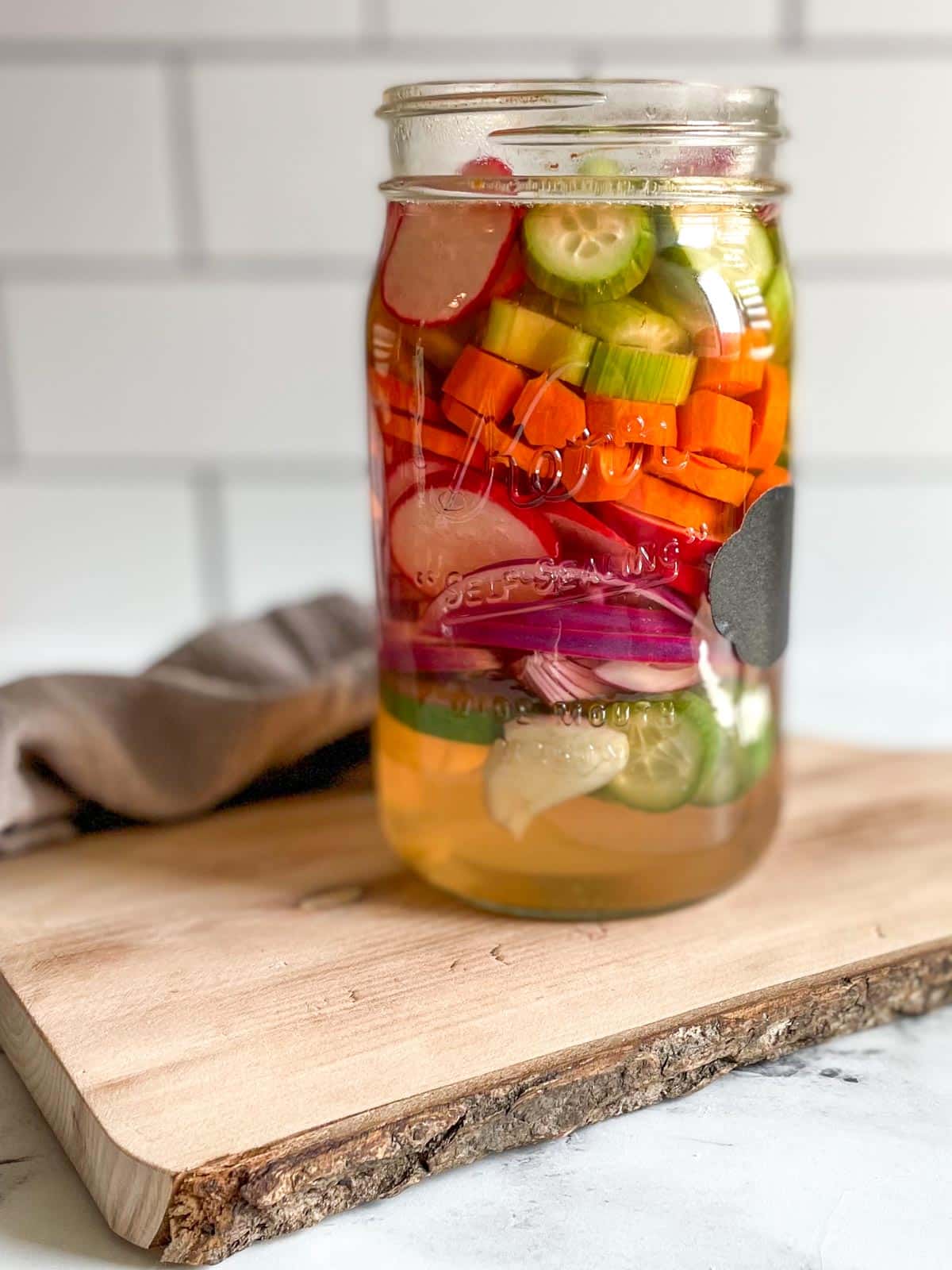 A glass jar of mixed pickles sits on a wooden cutting board with a gray linen.