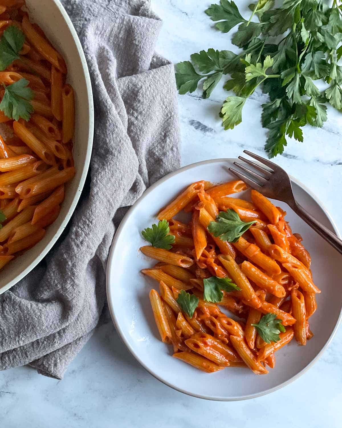 A white plate of Penne al Baffo garnished with parsley sits on a white marble counter with a gray linen, a fork, a frying pan full of penne al baffo, and a bunch of parsley.