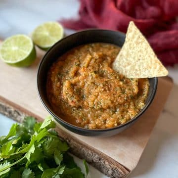 A bowl of pineapple habanero salsa sits on a rustic wooden cutting board surrounded by limes, cilantro, and a red linen.