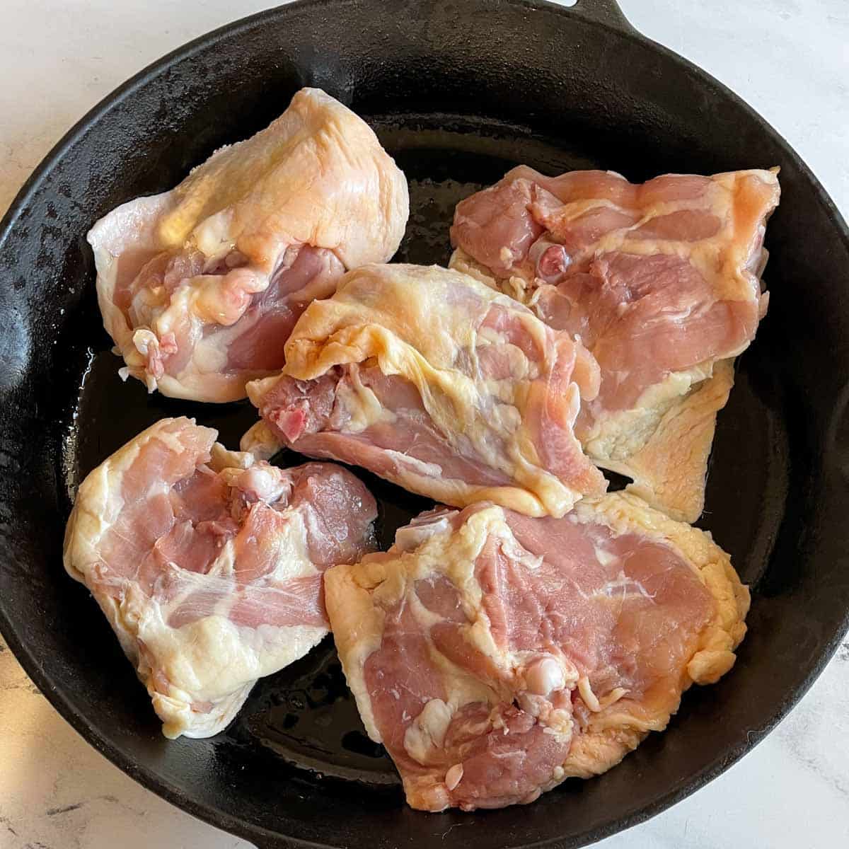 Five raw chicken thighs are shown skin side down in a black cast iron pan.